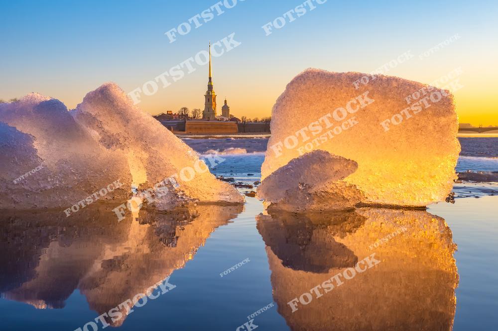 На переднем плане тающие льдины, которые плавают на реке Неве, отражая солнечные лучи. Вдали возвышается Петропавловская крепость с ее знаменитым шпилем, символизируя историческое величие Санкт-Петербурга. Теплый свет весеннего заката освещает воду и лед, создавая атмосферу пробуждения природы. Этот вид подчеркивает гармонию городской архитектуры и природных элементов.